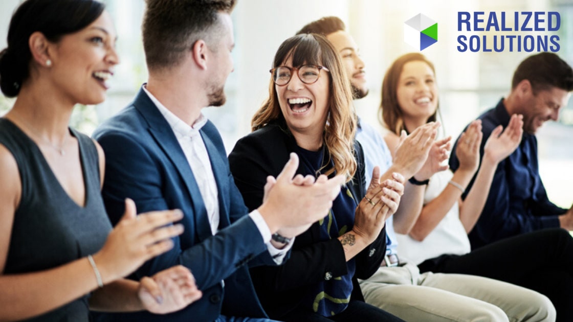 Group of professionals sitting in a row and applauding during a business event, with the Realized Solutions logo displayed in the upper right corner.