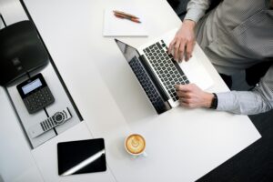 Person working on a laptop at a white office desk with a phone, tablet, notepad, and coffee.