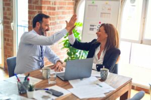 Business professionals celebrating a successful project with a high-five during a meeting, surrounded by reports, analytics, and a laptop in a modern office.