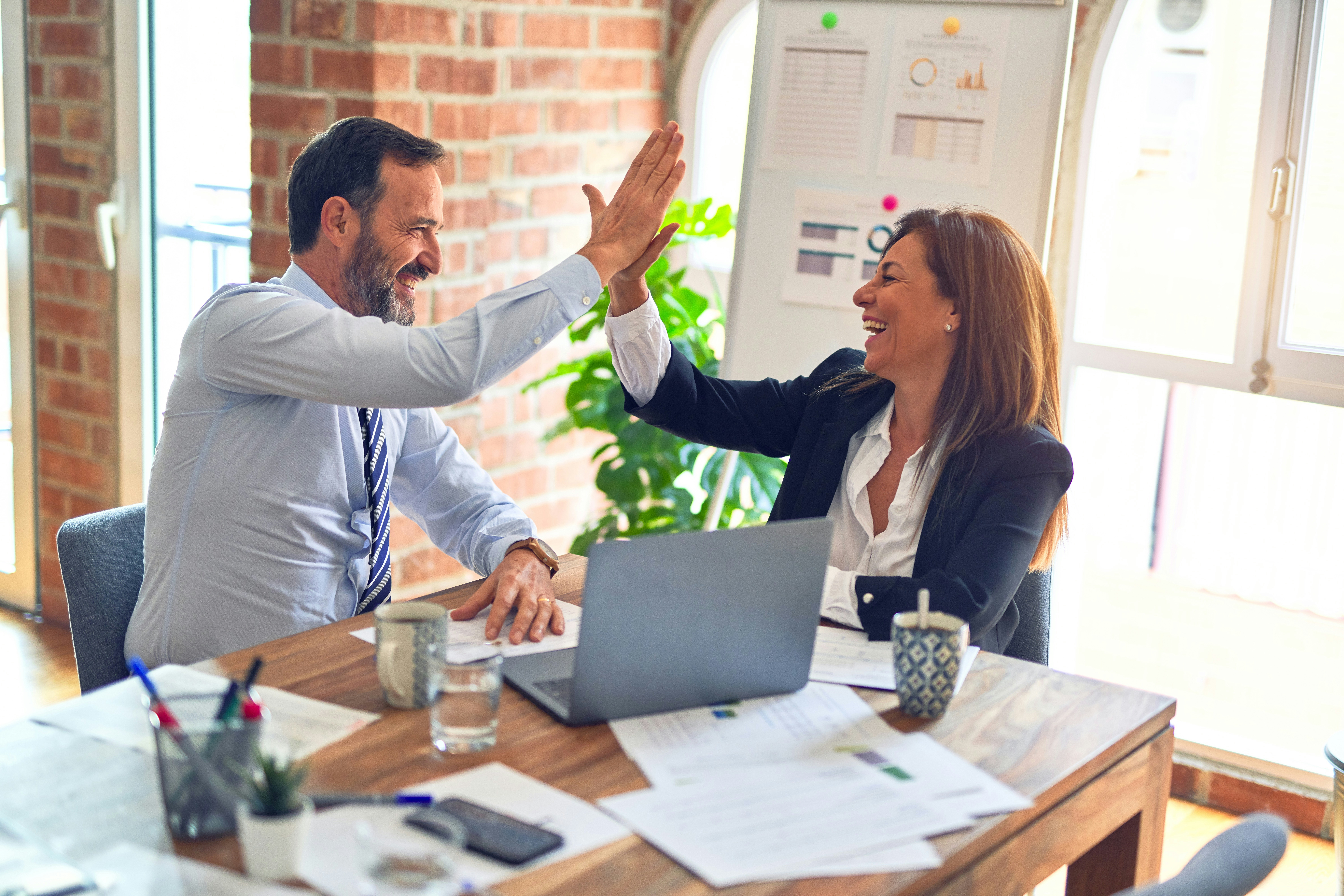 Business professionals celebrating a successful project with a high-five during a meeting, surrounded by reports, analytics, and a laptop in a modern office.