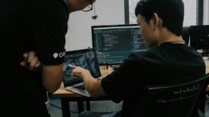 Software developers reviewing code on laptop and monitor screens while collaborating on programming and application development in an office setting.