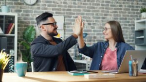 Two coworkers celebrating a successful project with a high-five at a desk in a modern office, highlighting teamwork, collaboration, and workplace success.