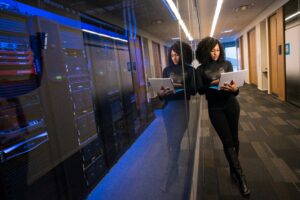 IT professional using a tablet in a data center hallway next to server racks, representing network management, cybersecurity, cloud infrastructure, and enterprise technology.