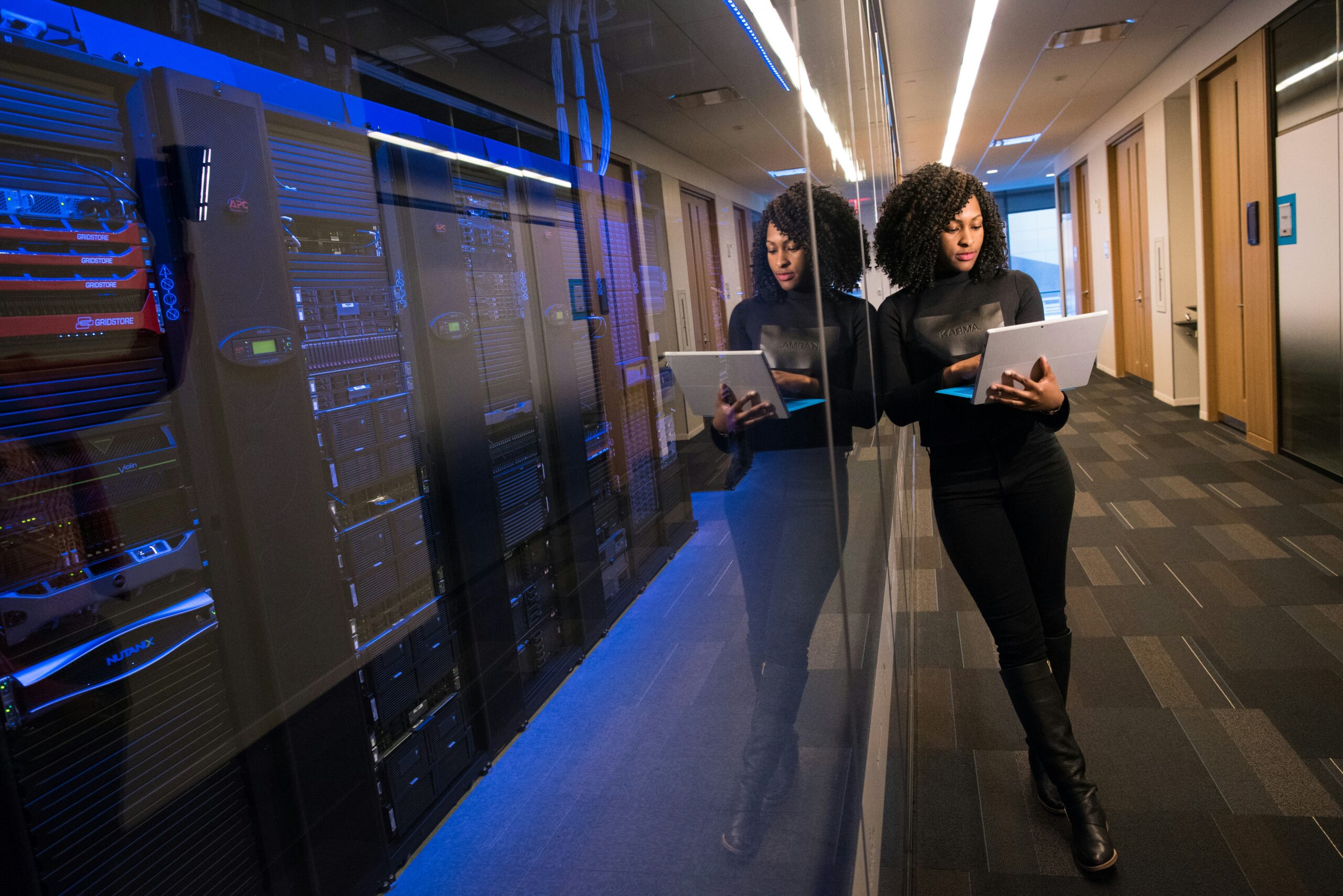 IT professional using a tablet in a data center hallway next to server racks, representing network management, cybersecurity, cloud infrastructure, and enterprise technology.