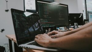 Software developer coding on a laptop and dual monitors displaying programming code in a professional workspace, representing application development and IT services.