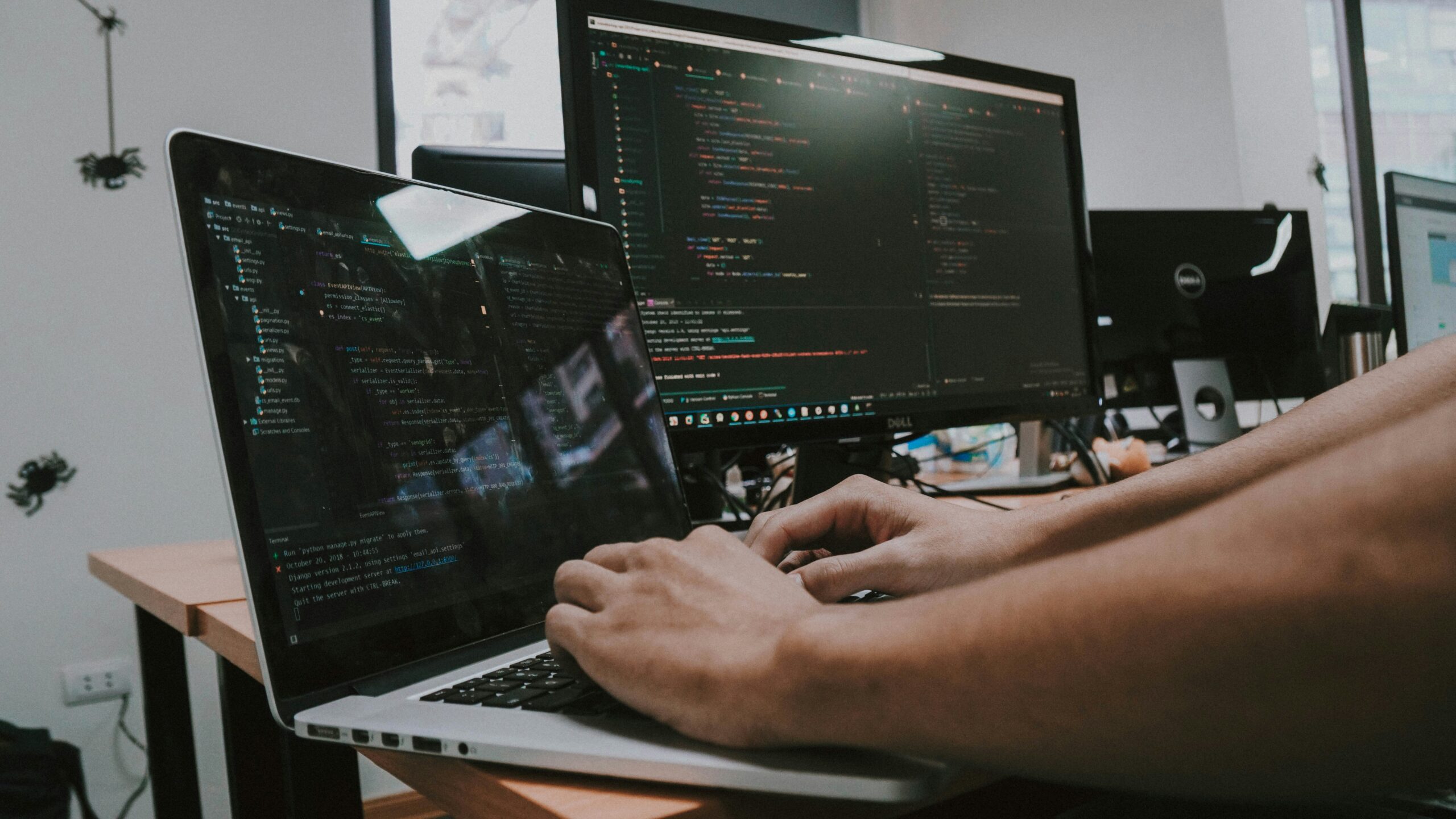 Software developer coding on a laptop and dual monitors displaying programming code in a professional workspace, representing application development and IT services.